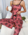 Woman in pink milk silk suspender pajamas with crown design, holding a cup, seated on a bed.